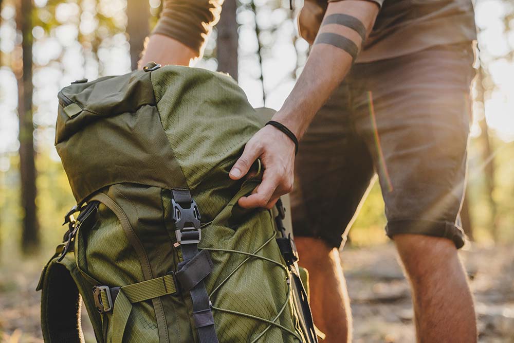 Tipps und Packliste für die Kanutour Person mit Arm-Tätowierung hält grünen Rucksack in einem sonnenbeschienenen Wald.