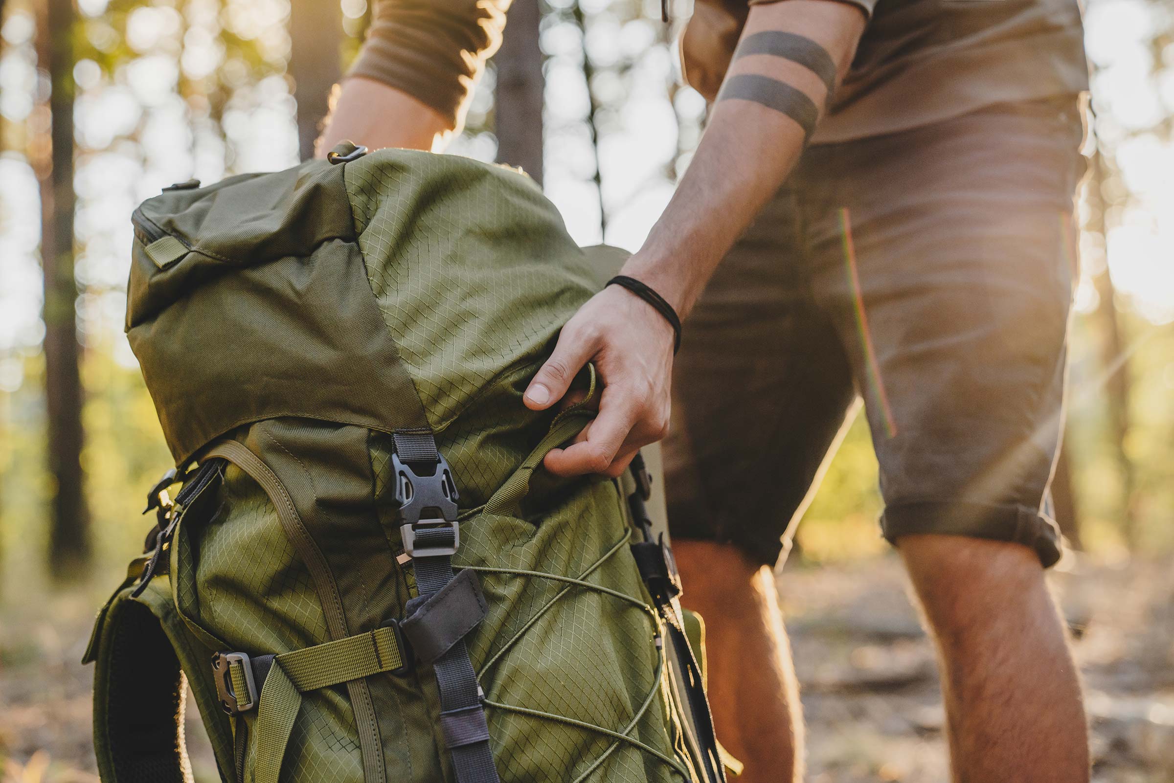 Jemand hält den Griff eines großen grünen Rucksacks, im Wald bei Sonnenlicht.