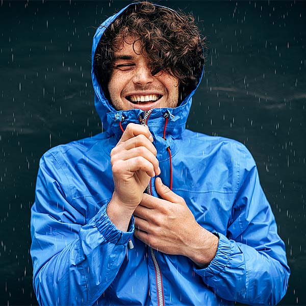 Mann im blauen Regenmantel mit Kapuze lächelt im Regen und zieht den Reißverschluss hoch.